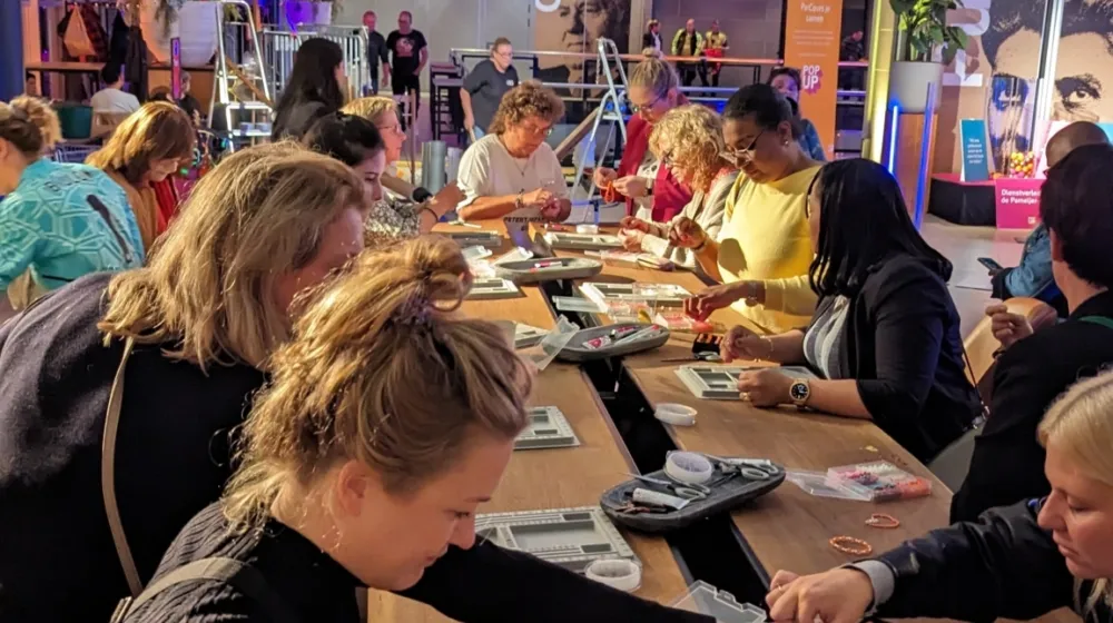 In een evenementenruimte staan lange tafels waaraan mensen samen armbanden maken