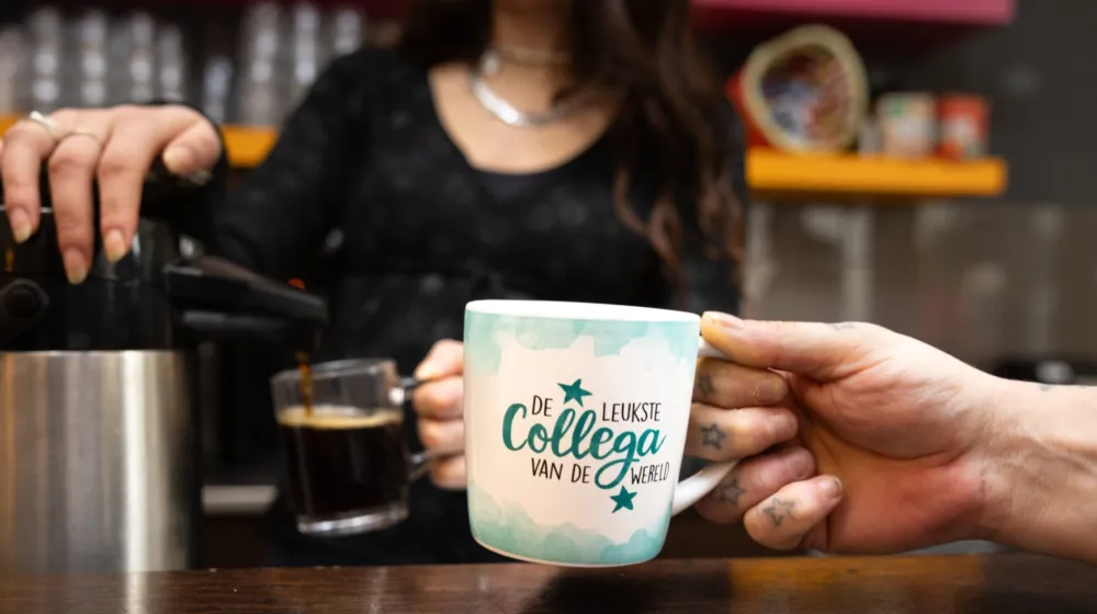 Vrouw schenkt koffie in en iemand heeft een mok met de leukste collega in de wereld mok vast