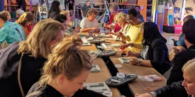 In een evenementenruimte staan lange tafels waaraan mensen samen armbanden maken