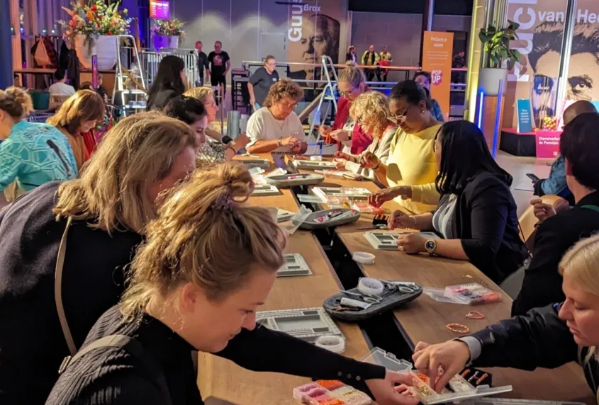 In een evenementenruimte staan lange tafels waaraan mensen samen armbanden maken