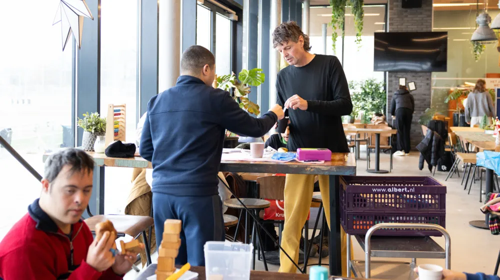 In een voetbalkantine staan twee mannen aan een tafel. Daar omheen staan meerdere tafels waar mensen aan zitten. Er staat eten en drinken op tafel.