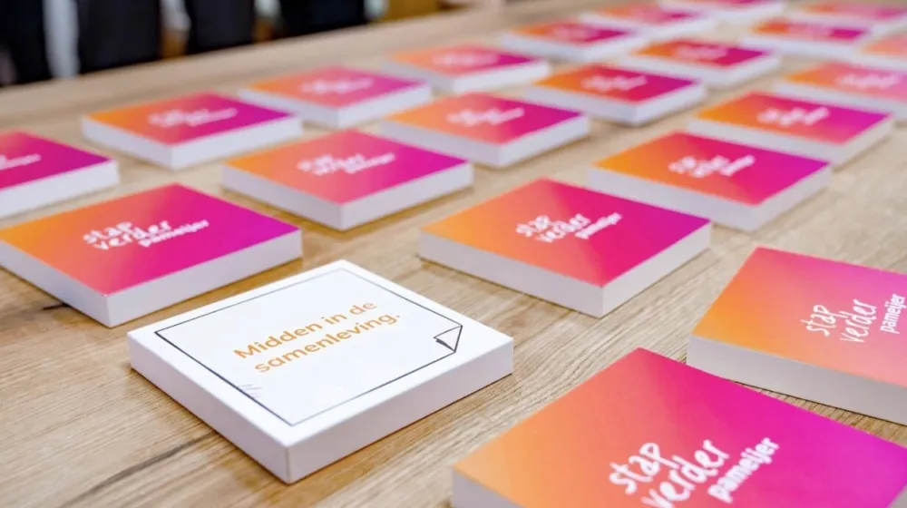 Op een tafel liggen vierkante plaatjes uitgestald. De roze plaatjes tonen het logo van Pameijer met de tekst 'Stap verder'. Eén wit plaatje bevat de tekst 'Midden in de samenleving'.