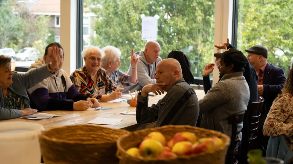 In een kantineruimte zitten aan een lange tafel een groep ouderen. Op de voorgrond staan twee manden met fruit.