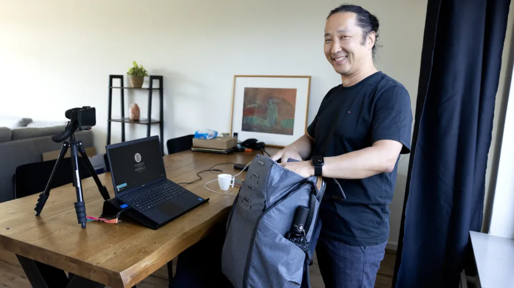 Een man staat in een woonkamer zijn rugtas uit te pakken. Zijn laptop en camera met statief staan op tafel.