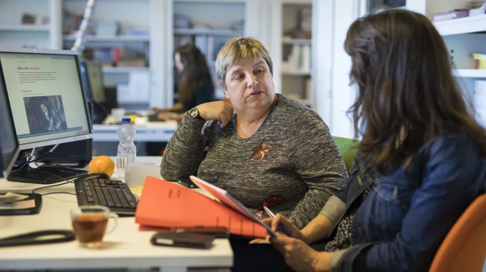 Twee vrouwen zitten in een kantoor ruimte achter een bureau. Ze zijn in gesprek met elkaar. Op het bureau staat een computer scherm aan en staat een kopje thee, een flesje water en een mandarijn.