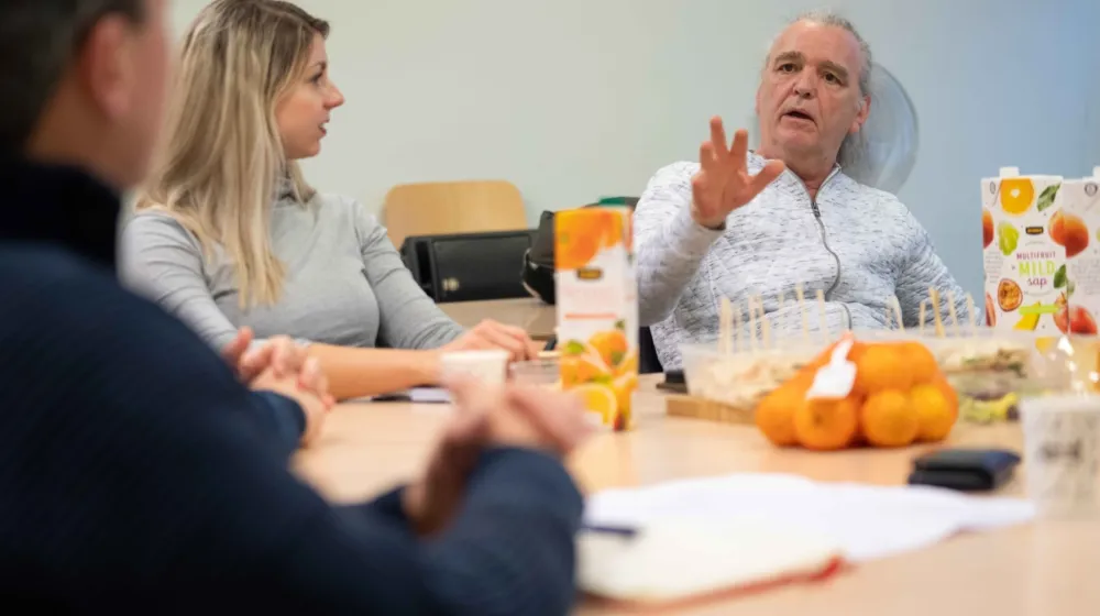 Een vrouw en twee mannen zitten aan een grote lunch tafel. Ze zijn in gesprek met elkaar. Op de tafel ligt een netje met mandarijnen en staan er drie pakken sap.