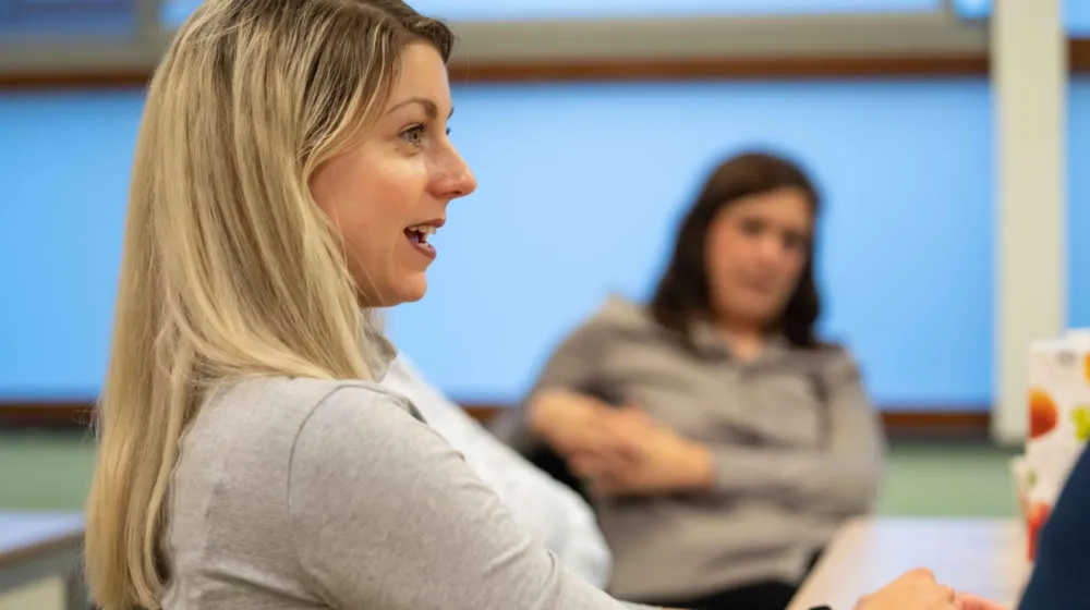 Een vrouw zit aan een tafel. De vrouw is in gesprek. Op de achtergrond zit een andere vrouw naar het gesprek te luisteren.
