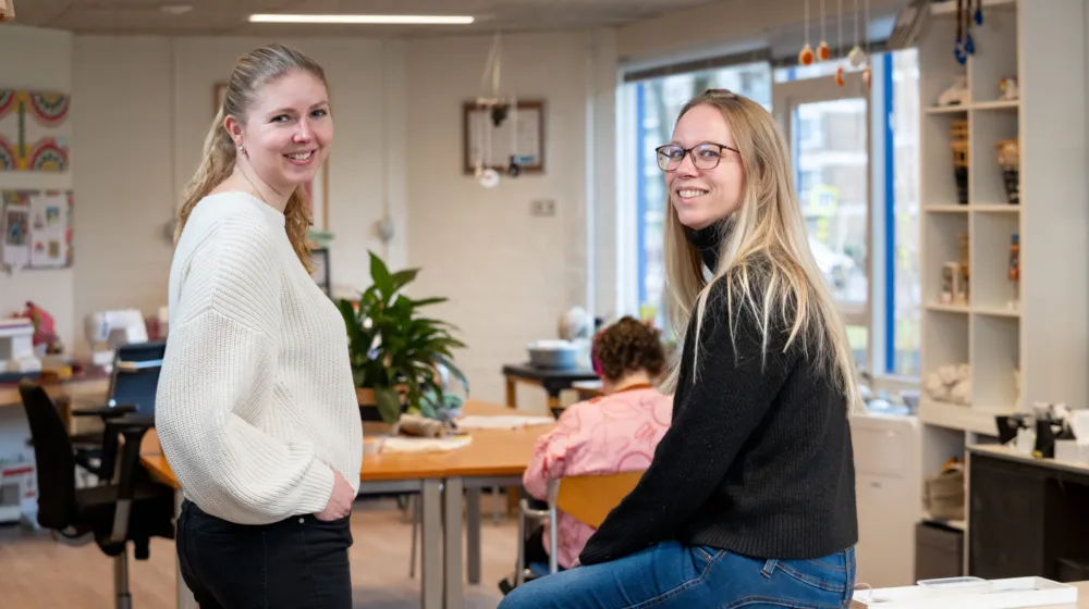 In een kantoor ruimte zit een vrouw op een bureau. Ze lacht naar de camera. Naast haar staat een vrouw. Zijn lacht ook naar de camera.