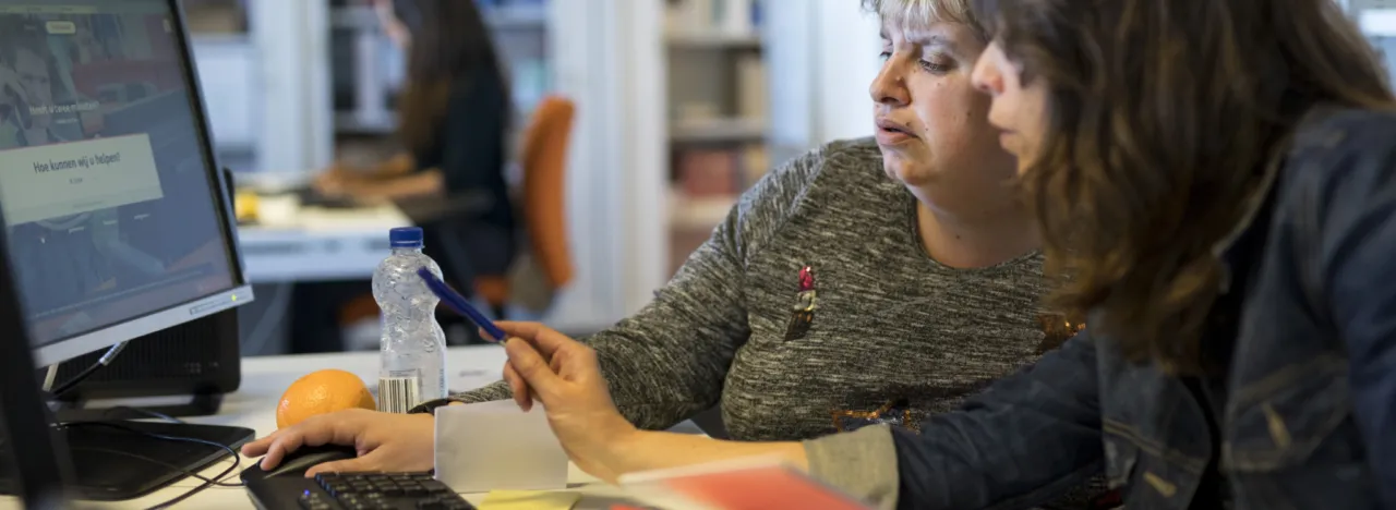 Twee vrouwen zitten aan een bureau. Ze kijken naar een computerscherm. De ene vrouw begeleid de andere vrouw waar ze met de computermuis moet klikken.