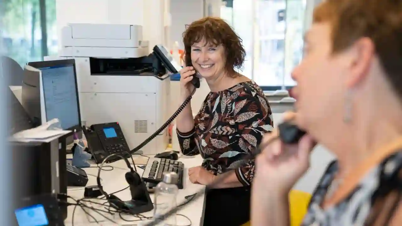 Twee vrouwen zitten achter de receptiebalie. Ze zijn aan het telefoneren. Ze kijken naar elkaar en glimlachen.