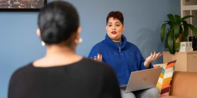 Twee vrouwen zitten tegen over elkaar te praten. Een heeft een laptop op haar schoot.