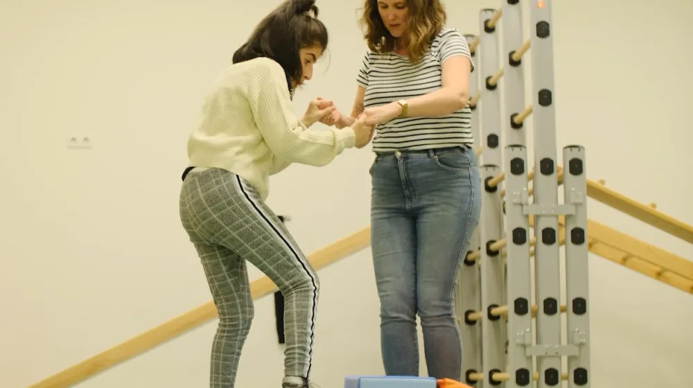 In een gymzaal houdt een vrouw de handen van een jonge vrouwelijke cliënt vast. De jonge vrouwelijke cliënt zet een stap op een kleine trap treden. Achter hen staat een klimrek.