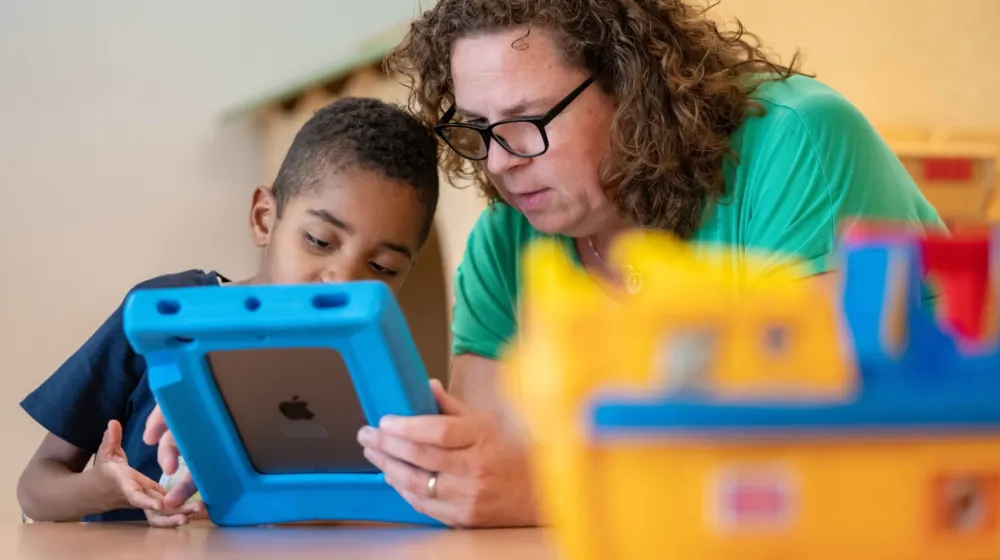 Een vrouw zit samen met een jongetje aan tafel. De vrouw houdt een Ipad vast en samen kijken ze naar het scherm. Op de voorgrond staat er speelgoed op tafel.