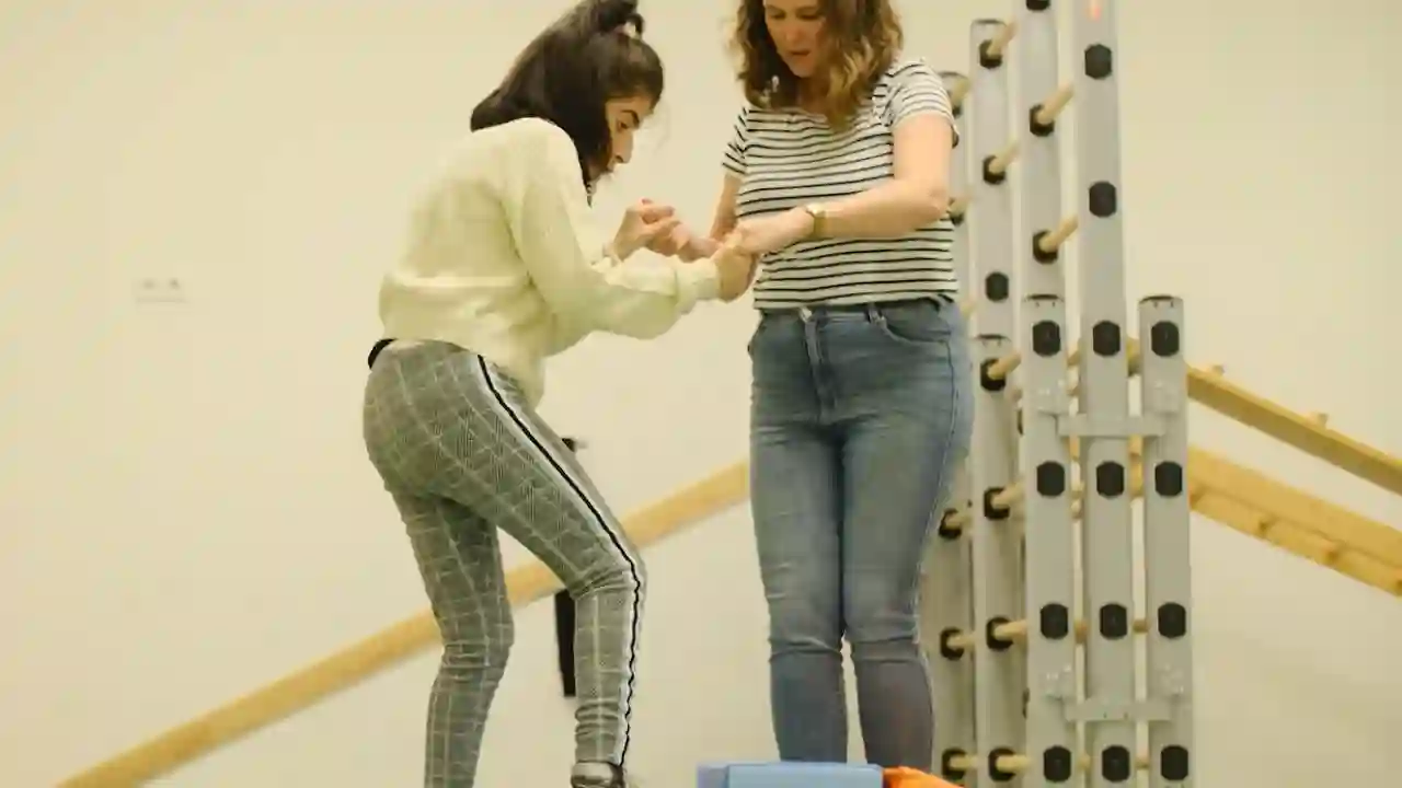In een gymzaal houdt een vrouw de handen van een jonge vrouwelijke cliënt vast. De jonge vrouwelijke cliënt zet een stap op een kleine trap treden. Achter hen staat een klimrek.