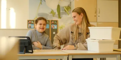 In een klaslokaal zitten een meisje en een vrouwelijke medewerker achter een bureautje te lachen. Op het bureautje staat een Ipad en wat witte bakken.