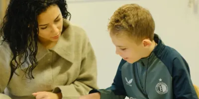 Een jongetje en vrouw zitten aan tafel. Het jongetje wijst naar een plaat op tafel en de vrouw kijkt naar de plaat.