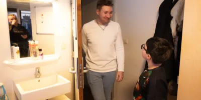 Een jongetje staat in een badkamer bij een gootsteen met een spiegel. In de deur opening staat een man die naar het jongetje glimlacht.