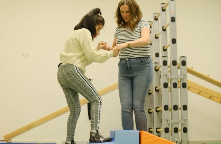 In een gymzaal houdt een vrouw de handen van een jonge vrouwelijke cliënt vast. De jonge vrouwelijke cliënt zet een stap op een kleine trap treden. Achter hen staat een klimrek.
