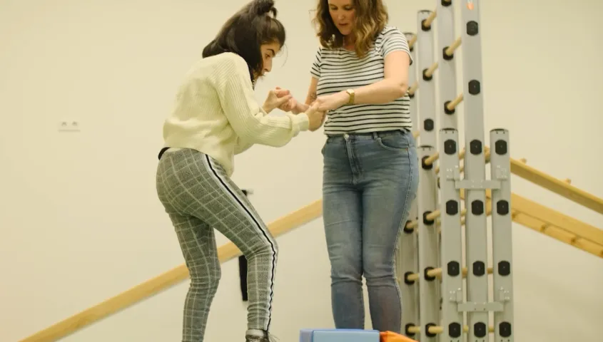 In een gymzaal houdt een vrouw de handen van een jonge vrouwelijke cliënt vast. De jonge vrouwelijke cliënt zet een stap op een kleine trap treden. Achter hen staat een klimrek.