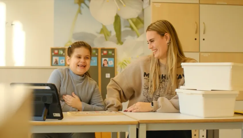 In een klaslokaal zitten een meisje en een vrouwelijke medewerker achter een bureautje te lachen. Op het bureautje staat een Ipad en wat witte bakken.