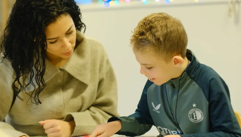 Een jongetje en vrouw zitten aan tafel. Het jongetje wijst naar een plaat op tafel en de vrouw kijkt naar de plaat.
