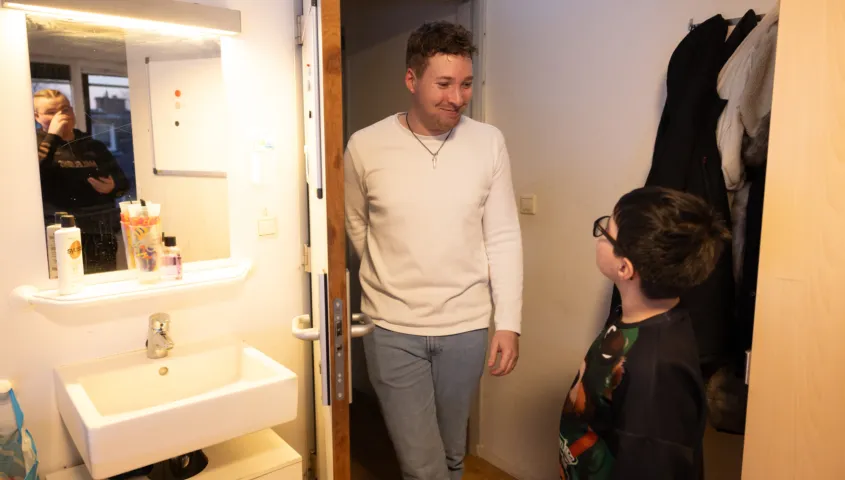 Een jongetje staat in een badkamer bij een gootsteen met een spiegel. In de deur opening staat een man die naar het jongetje glimlacht.