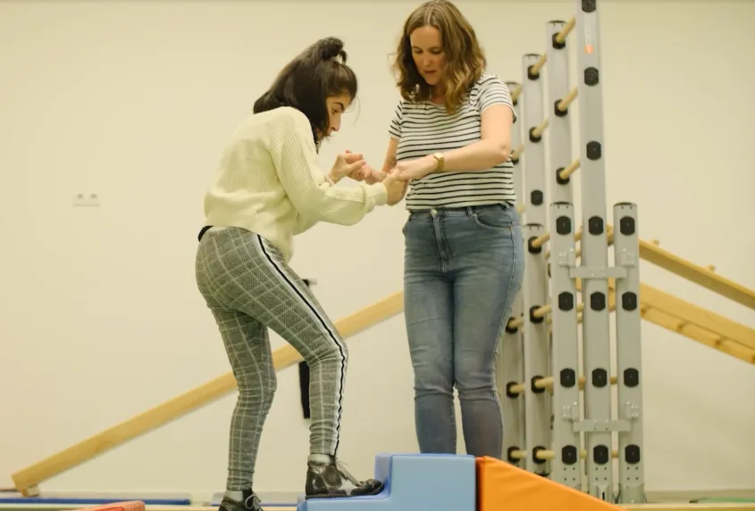 In een gymzaal houdt een vrouw de handen van een jonge vrouwelijke cliënt vast. De jonge vrouwelijke cliënt zet een stap op een kleine trap treden. Achter hen staat een klimrek.