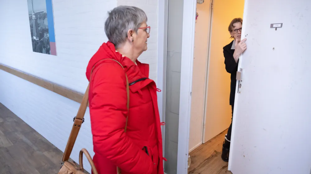Een vrouw staat in een witte hal voor de deur van een vrouwelijke cliënt. De vrouwelijke cliënt opent de deur.