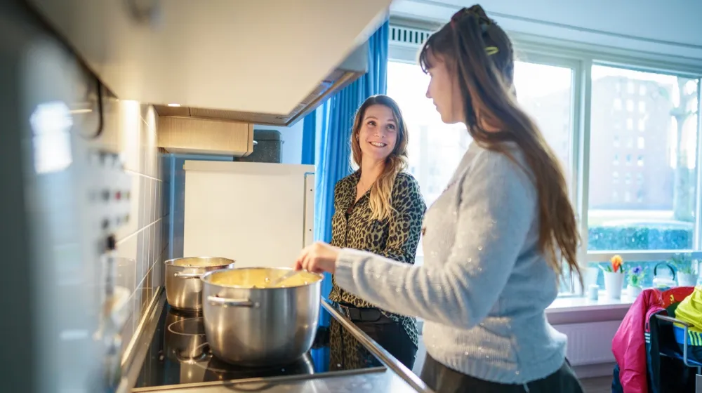 Twee vrouwen staan in de keuken achter het fornuis. Er staan twee pannen op het fornuis met aardappelen. De vrouwen glimlachen naar elkaar.