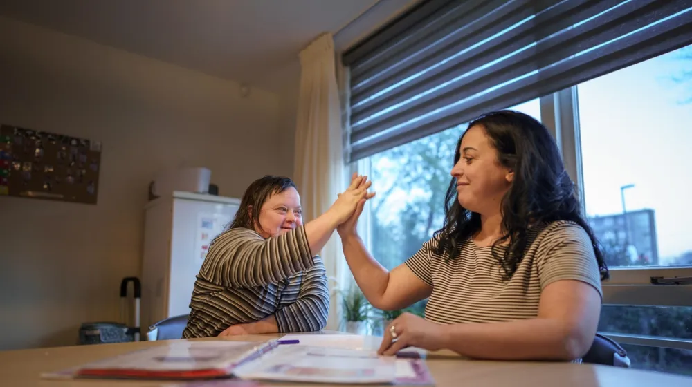 Een vrouw zit met een vrouwelijke cliënt aan tafel. Ze glimlachen naar elkaar en geven een high five. Op tafel ligt een map open met documenten.