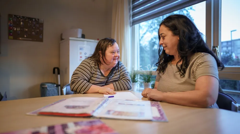 Een vrouw zit met een vrouwelijke cliënt aan tafel in een woonkamer. Ze glimlachen naar elkaar. Op tafel ligt een map open met wat documenten erin.