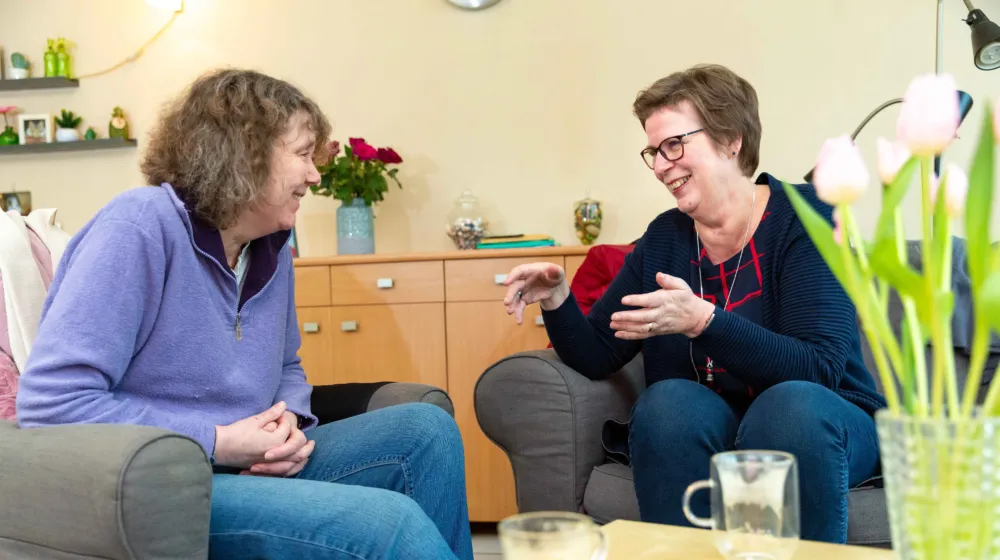 Twee vrouwen zitten in een woonkamer aan de koffie tafel. Ze glimlachen naar elkaar en zijn in gesprek. Op tafel staan twee koffie kopjes en een vaas met witte tulpen.
