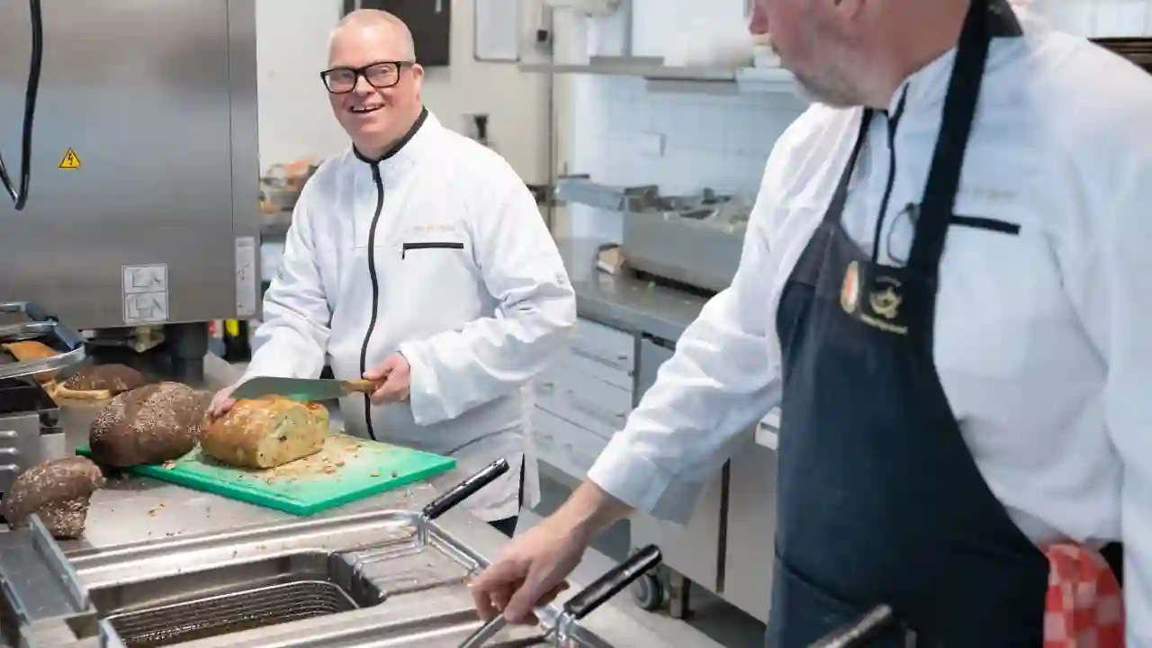 Twee mannen staan in een restaurant keuken. Een man is brood aan het snijden. Hij lacht naar de andere man die aan het frituren is in een frituurpan.