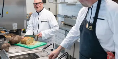 Twee mannen staan in een restaurant keuken. Een man is brood aan het snijden. Hij lacht naar de andere man die aan het frituren is in een frituurpan.