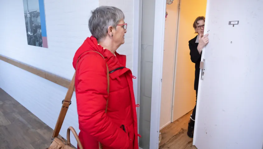Een vrouw staat in een witte hal voor de deur van een vrouwelijke cliënt. De vrouwelijke cliënt opent de deur.