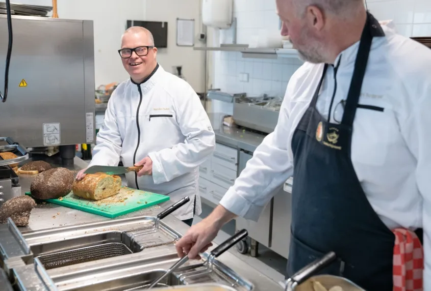 Twee mannen staan in een restaurant keuken. Een man is brood aan het snijden. Hij lacht naar de andere man die aan het frituren is in een frituurpan.