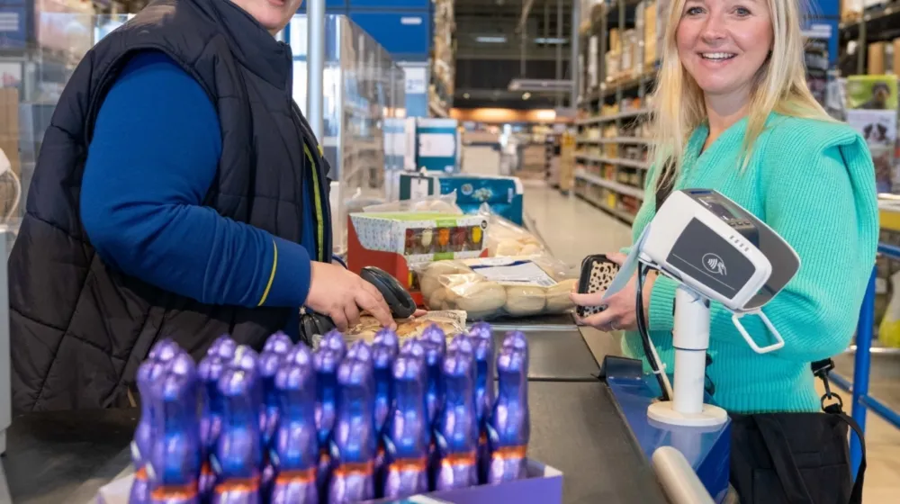 Een vrouw zit achter de kassa van een supermarkt. Een vrouwelijke klant staat aan de kassa terwijl haar boodschappen worden gescand.
