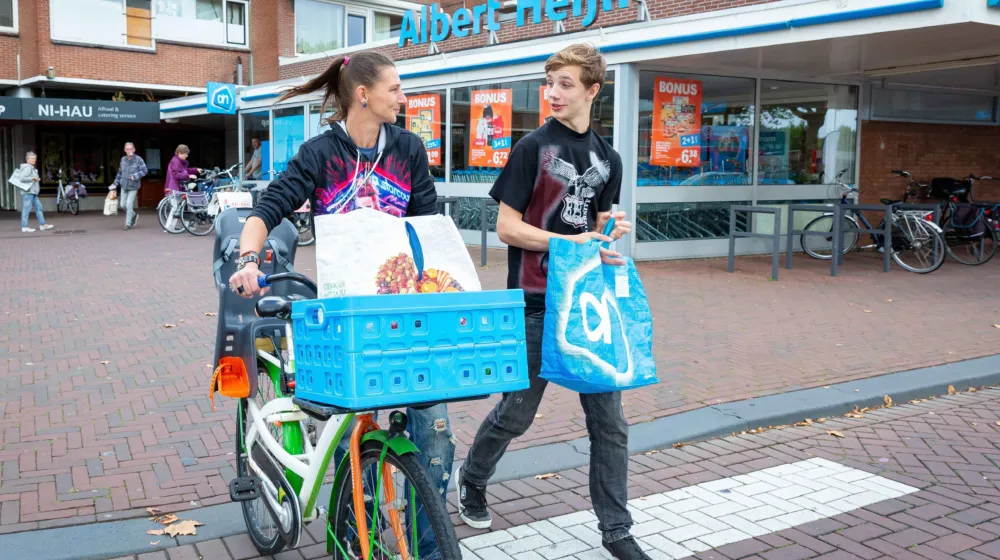 Buiten op straat loopt een vrouw met een fiets in haar hand en een boodschappentas in de fietsmand. Naast haar loopt een jongen met een boodschappentas in zijn handen. Op de achtergrond is de supermarkt.
