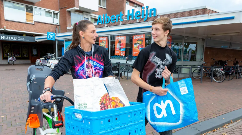 Op straat loopt een vrouw met haar fiets in de hand en een boodschappentas in de mand voorop. Naast haar loopt een jongen met een boodschappentas in zijn hand. Op de achtergrond is een supermarkt.