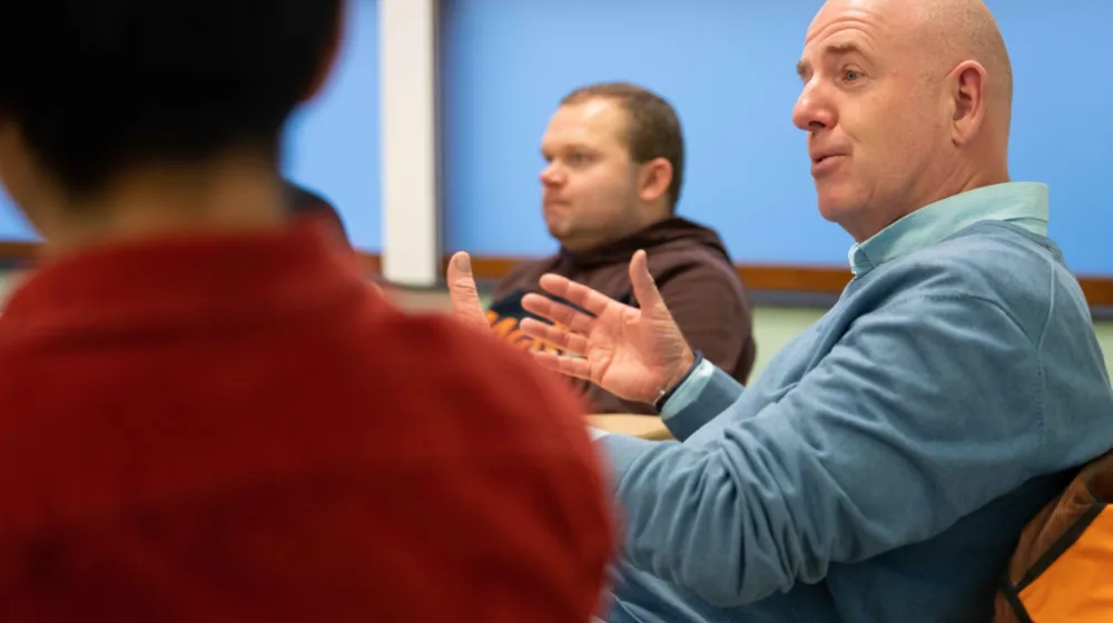 Drie mannen zitten in een kantoor ruimte aan een tafel. Ze zijn in gesprek met elkaar.