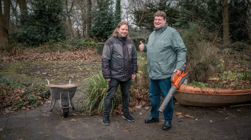 Twee mannen staan buiten in een grote tuin met op de achtergrond bomen. Het is herfst en de bladeren liggen op de grond. Ze lachen beide naar de camera en één man doet zijn duim omhoog. Hij heeft een bladblazer vast. Links van de mannen staat een kruiwagen.