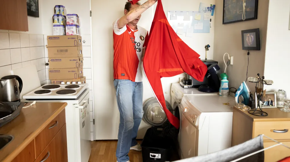 In een leefruimte staat een man bij de wasmachine en trekt hij een shirt uit de was. Achter hem staat een keukenblad met een fornuis.