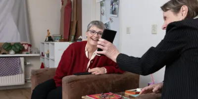 Twee vrouwen zitten in een woonkamer op een stoel. Een vrouw laat de andere vrouw haar mobiel zien. Ze lachen beide terwijl ze naar de mobiel kijken.