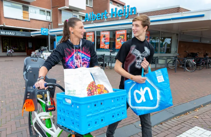 Op straat loopt een vrouw met haar fiets in de hand en een boodschappentas in de mand voorop. Naast haar loopt een jongen met een boodschappentas in zijn hand. Op de achtergrond is een supermarkt.