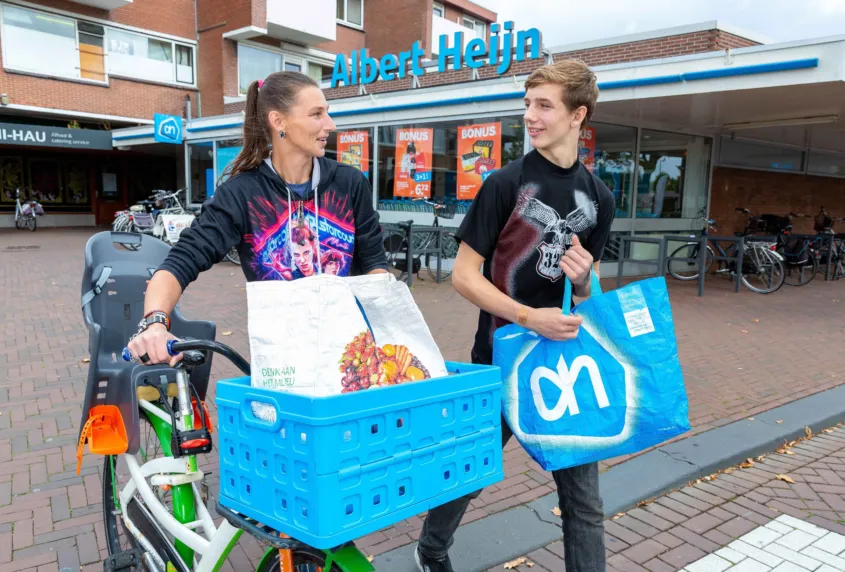 Op straat loopt een vrouw met haar fiets in de hand en een boodschappentas in de mand voorop. Naast haar loopt een jongen met een boodschappentas in zijn hand. Op de achtergrond is een supermarkt.